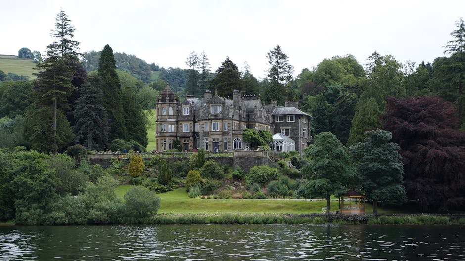 The image showcases a large, elegant residential property with a cream-colored facade and a red-tiled roof, situated behind a landscaped garden. In the foreground, there are ornamental grasses and roc