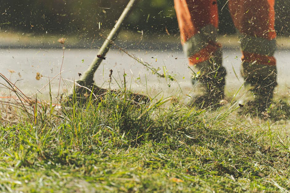 Close-up view of a person wearing orange waterproof gardening trousers and sturdy boots using a rake to clear away grass cuttings and weeds from a garden bed with overgrown grass, weeds, and scattered