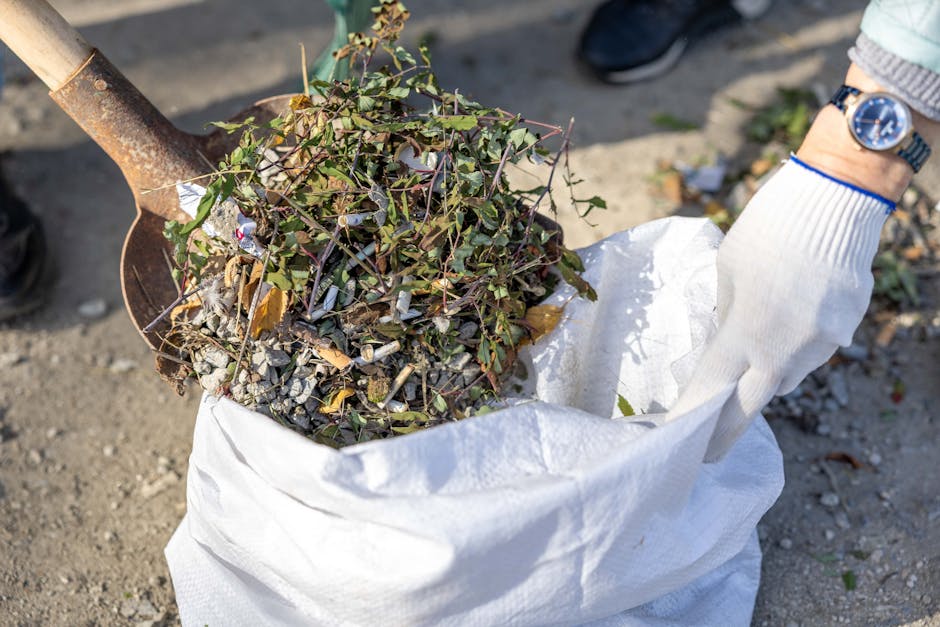 Close-up view of a person wearing orange waterproof gardening trousers and sturdy boots using a rake to clear away grass cuttings and weeds from a garden bed with overgrown grass, weeds, and scattered